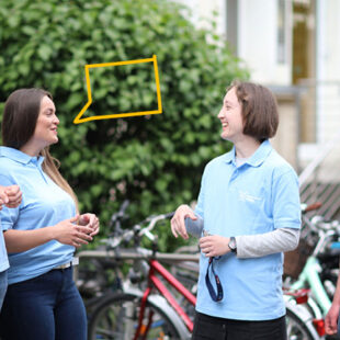 Vier Studierende mit einheitlichen hellblauen Shirts stehen vor einem grünen Hintergrund und unterhalten sich