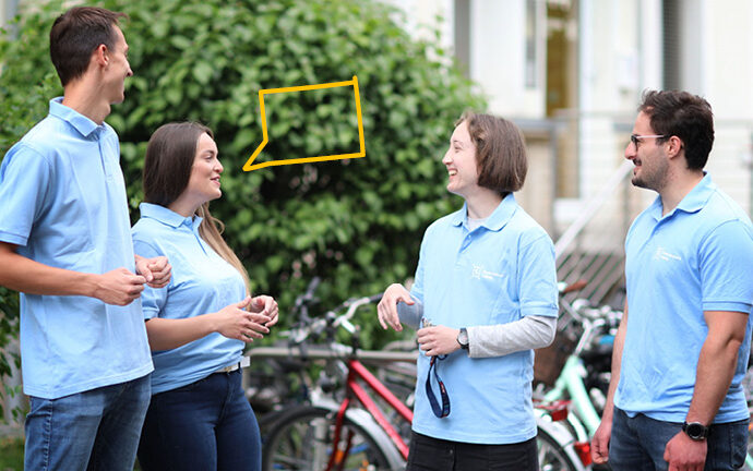 Vier Studierende mit einheitlichen hellblauen Shirts stehen vor einem grünen Hintergrund und unterhalten sich