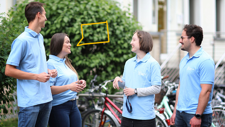 Vier Studierende mit einheitlichen hellblauen Shirts stehen vor einem grünen Hintergrund und unterhalten sich