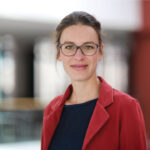 Frau mit Brille und roter Jacke lächelt in die Kamera.