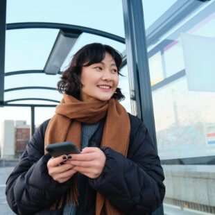 Eine junge Frau steht mit einem Handy in der Hand an einer Haltstelle und lächelt.