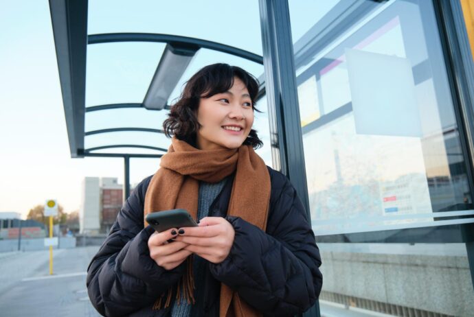 Eine junge Frau steht mit einem Handy in der Hand an einer Haltstelle und lächelt.