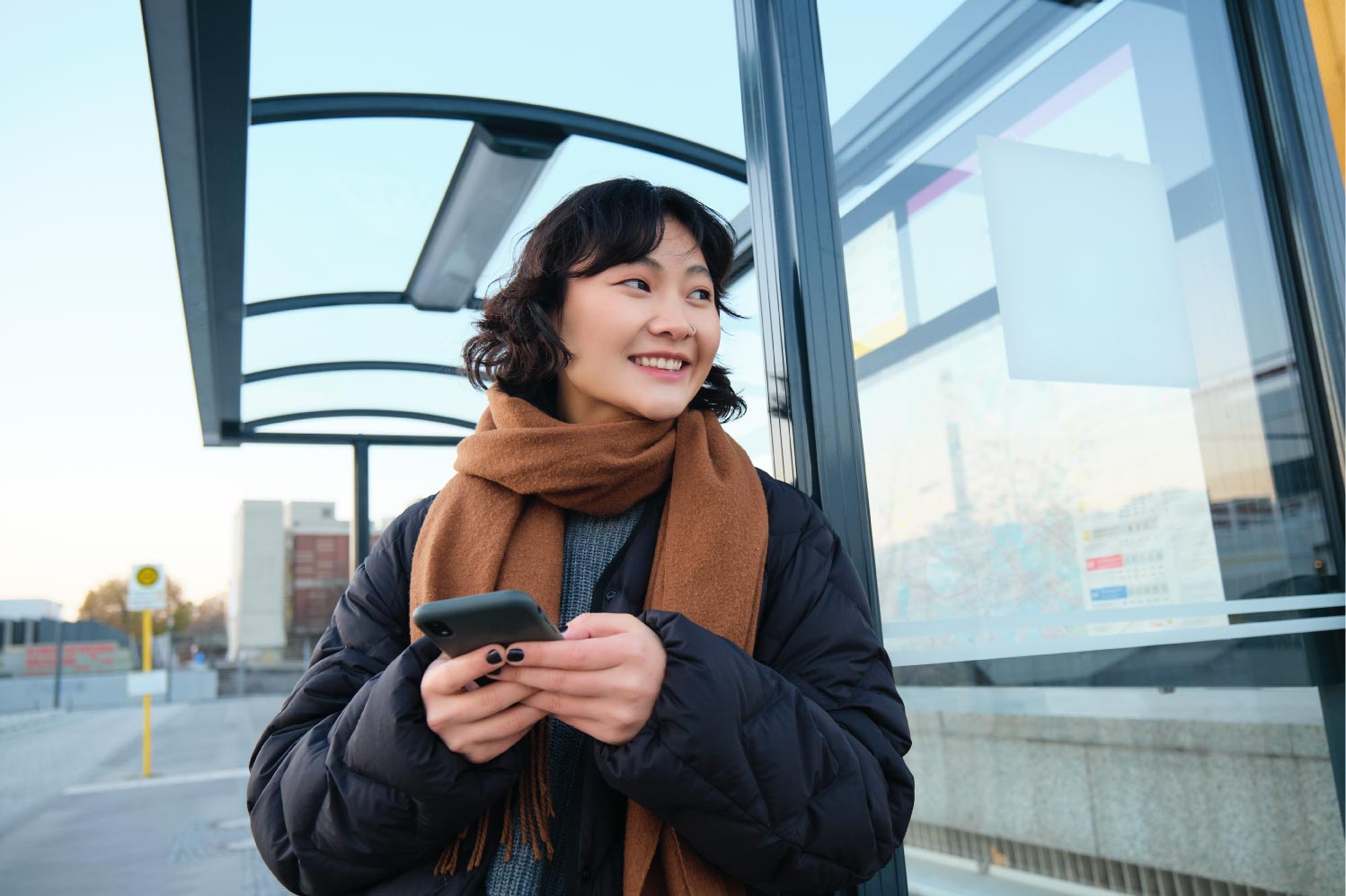 Eine junge Frau steht mit einem Handy in der Hand an einer Haltstelle und lächelt.