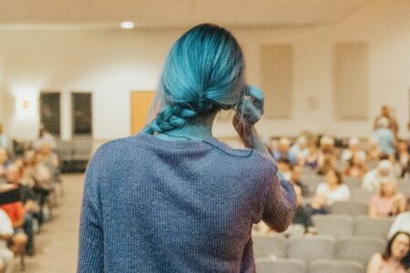 Eine Frau steht vor Menschen in einer AArt Hörsaal. Sie fasst sich nervös ans Ohr. Man sieht sie von hinten.