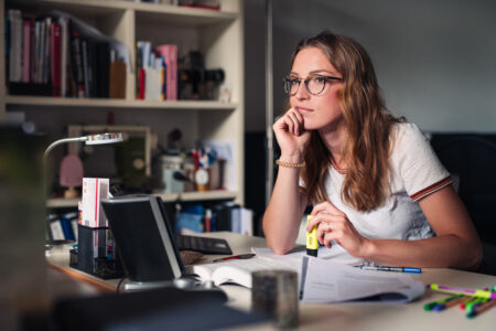 Eine junge Frau sitzt am Schreibtisch und erledigt Textarbeit für ihr Studium. Sie schaut sehr nachdenklich.