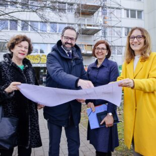 Dr. Beate Schücking (DSW-Präsidentin), Sebastian Gemkow (Wissenschaftsminister Sachsen), Dr. Andrea Diekhof (Geschäftsführerin des Studentenwerkes Leipzig) und Monika Schwarzenberg (Leiterin Studentisches Wohnen Studentenwerk Leipzig) stehen vor dem Wohnheim und halten einen Bauplan in den Händen. Dabei lächeln sie in die Kamera.