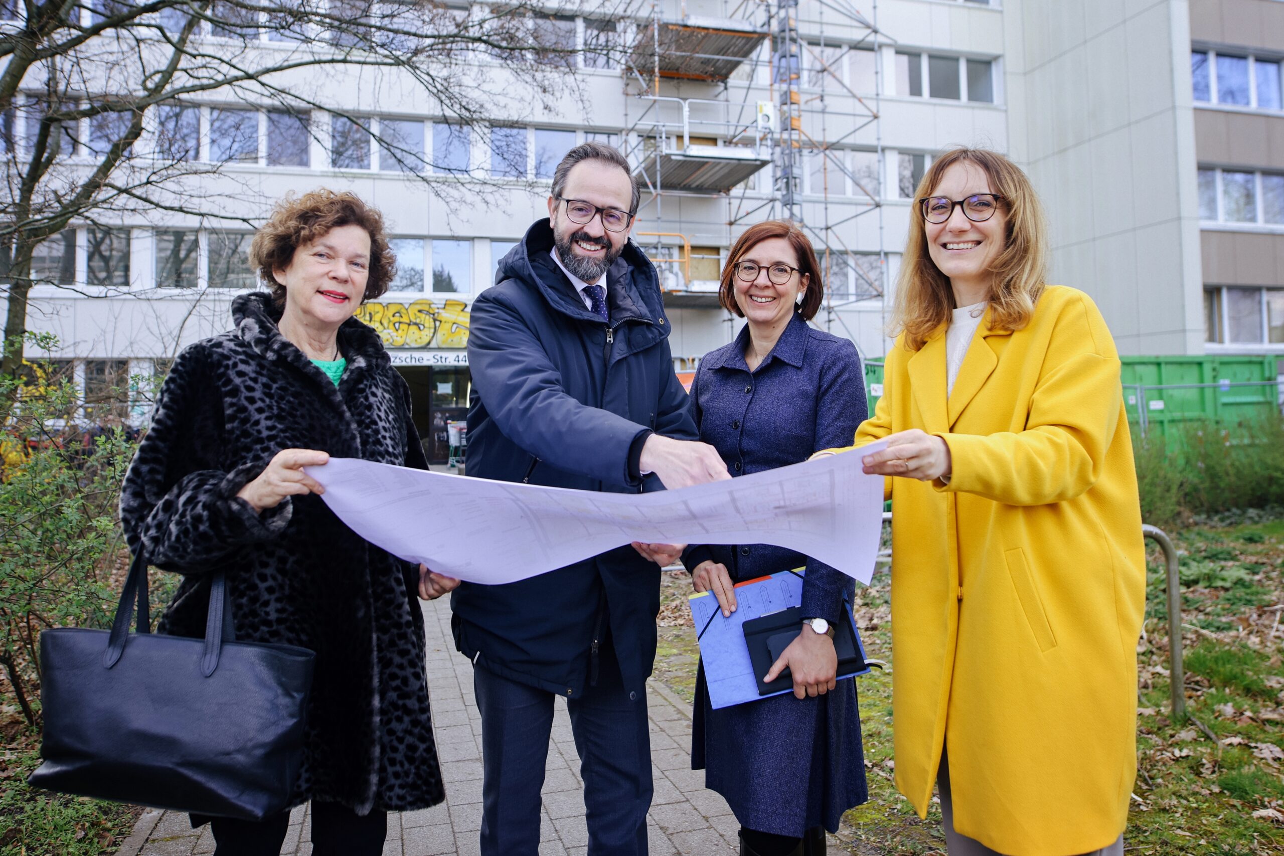 Dr. Beate Schücking (DSW-Präsidentin), Sebastian Gemkow (Wissenschaftsminister Sachsen), Dr. Andrea Diekhof (Geschäftsführerin des Studentenwerkes Leipzig) und Monika Schwarzenberg (Leiterin Studentisches Wohnen Studentenwerk Leipzig) stehen vor dem Wohnheim und halten einen Bauplan in den Händen. Dabei lächeln sie in die Kamera.