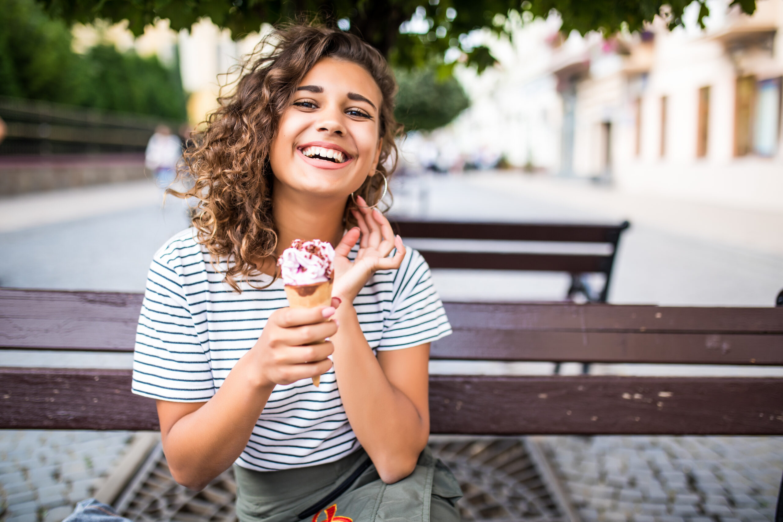 Frau sitzt auf einer Parkbank im Sommer und isst Eis, während sie in die Kamera lacht.