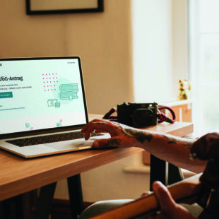 Junge Person sitzt am Schreibtisch und arbeitet am Laptop. Die BAföG Digital Website ist offen.