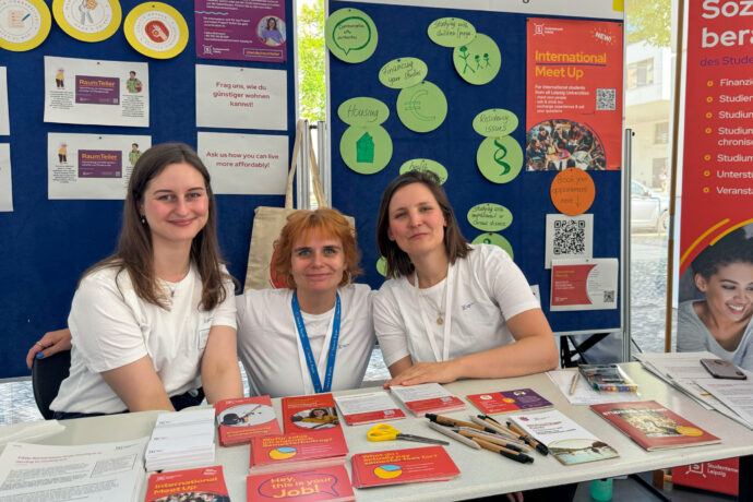 Drei Mitarbeiterinnen des Studentenwerks sitzen an ihrem Informationsstand und lächeln in die Kamera.