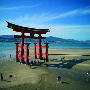 Rotes Torii-Tor auf Miyajima bei Ebbe, Menschen spazieren am Strand, Meer und Berge im Hintergrund.