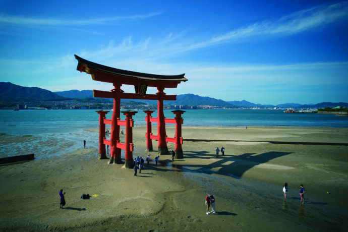 Rotes Torii-Tor auf Miyajima bei Ebbe, Menschen spazieren am Strand, Meer und Berge im Hintergrund.