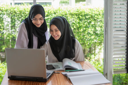 „Zwei junge Frauen mit schwarzen Kopftüchern sitzen gemeinsam an einem Tisch vor einem Laptop und lächeln.