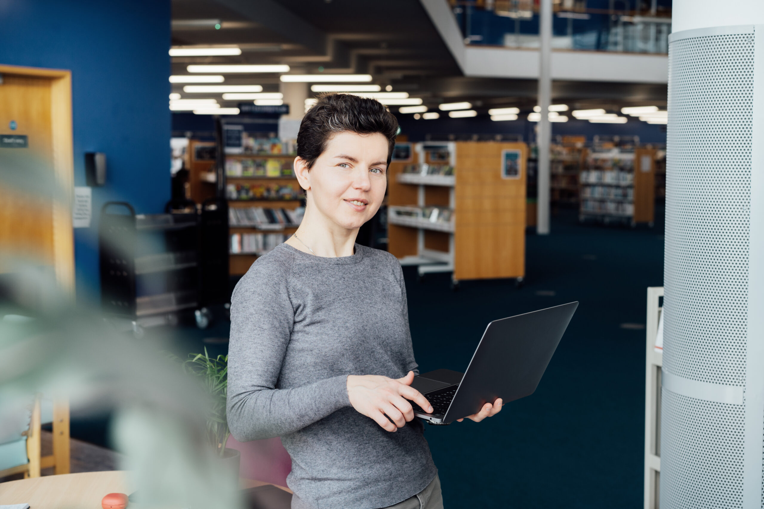 A person in a grey jumper holds a laptop in a library.