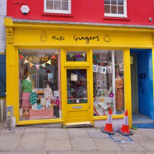 Bunte Fassade eines kleinen Ladens mit handgeschriebenem Schild „Miss Gingers“ und dekorierten Schaufenstern; zwei Verkehrshütchen davor.