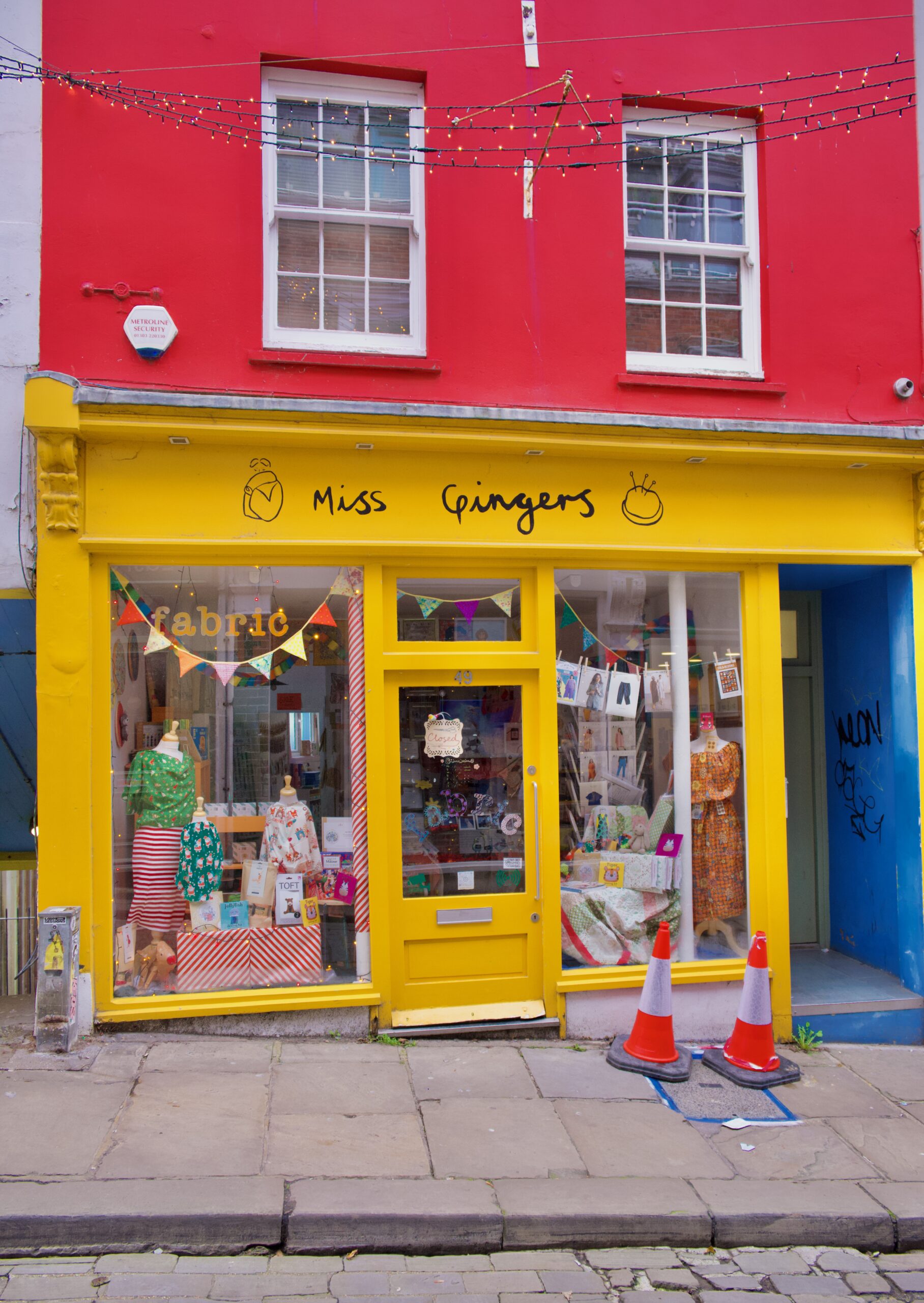 Bunte Fassade eines kleinen Ladens mit handgeschriebenem Schild „Miss Gingers“ und dekorierten Schaufenstern; zwei Verkehrshütchen davor.