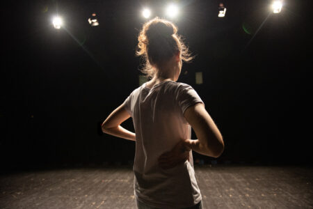Eine Person steht mit dem Rücken zur Kamera auf einer dunklen Bühne und hat die Hände in die Hüften gestützt. Mehrere helle Scheinwerfer leuchten von oben auf die Szene und erzeugen starke Lichtreflexe.