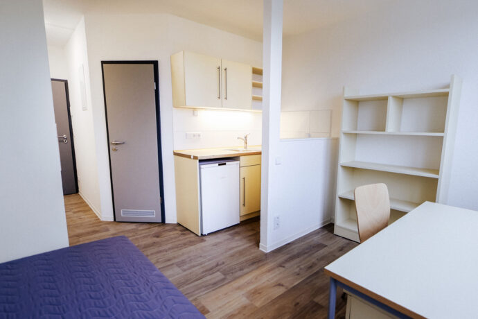 Studentenwohnheim mit Blick auf Eingangsbereich, Küche, Regale, Schreibtisch und Bett