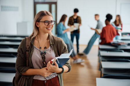 Eine Studentin hält Bücher in der Hand. Im Hintergrund sind andere Studierende.
