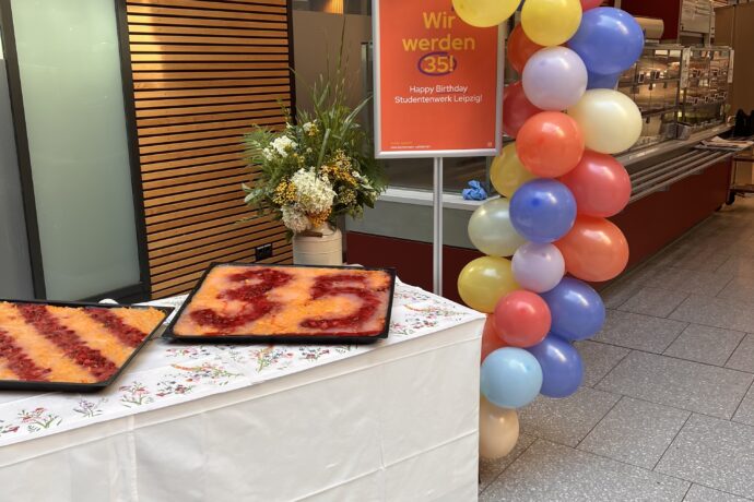 Zu sehen ist ein langer Tisch mit Obstkuchen auf Blechen. Daneben eine Traube Luftballons und ein Jubiläumsplakat.