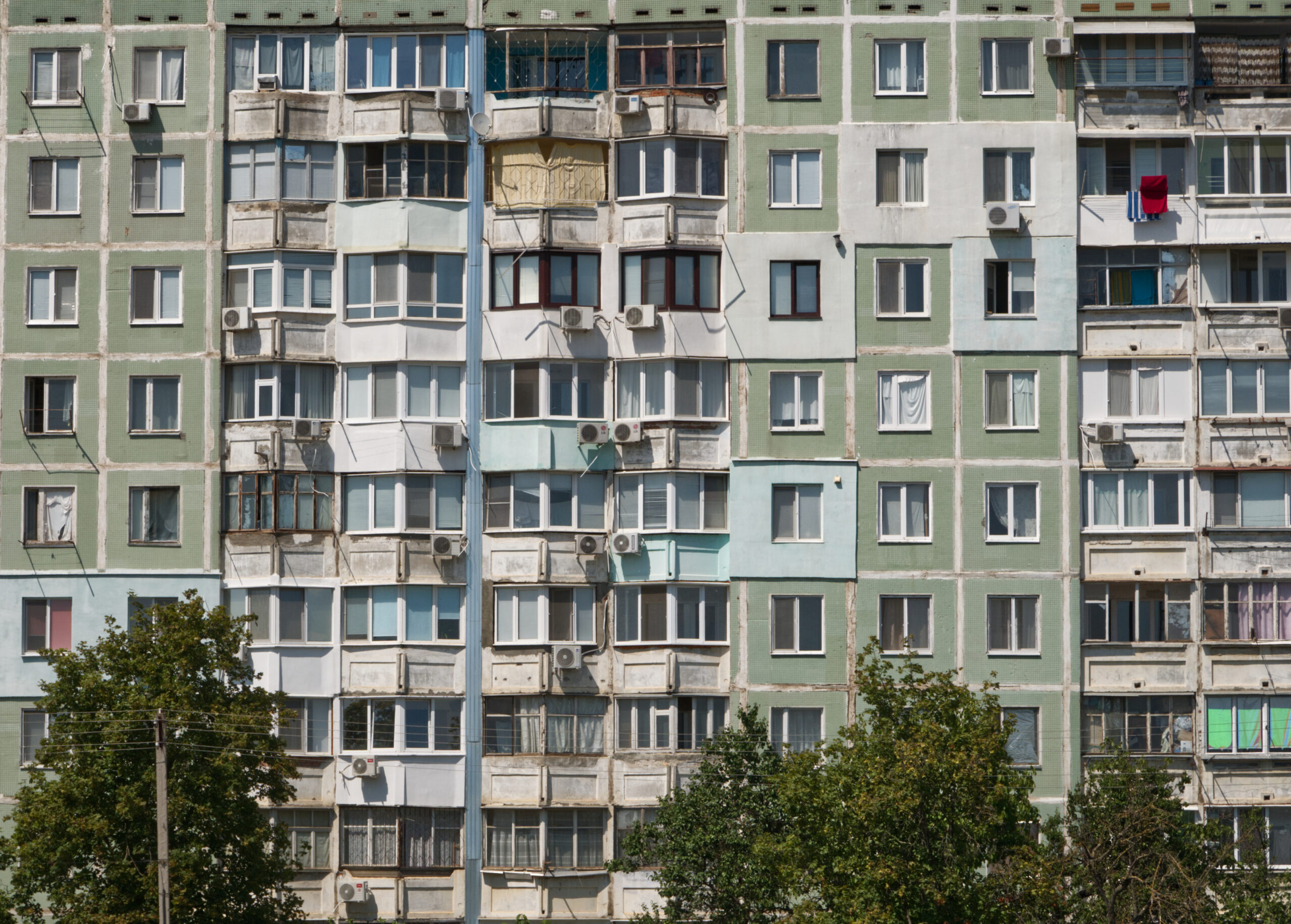 Mehrfamilienhaus mit Balkonen, Klimaanlagen und Bäumen im Vordergrund.