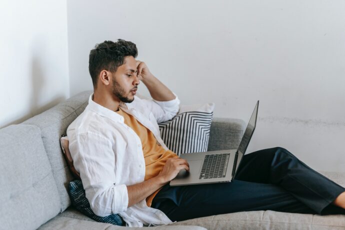 ein junger Mann sitzt mit Laptop auf dem Schoß auf dem Sofa und sieht nachdenklich aus