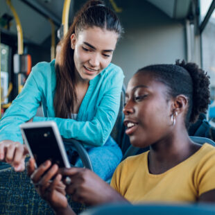 Zwei Frauen lachen gemeinsam und nutzen auf einer Busfahrt ein Smartphone.