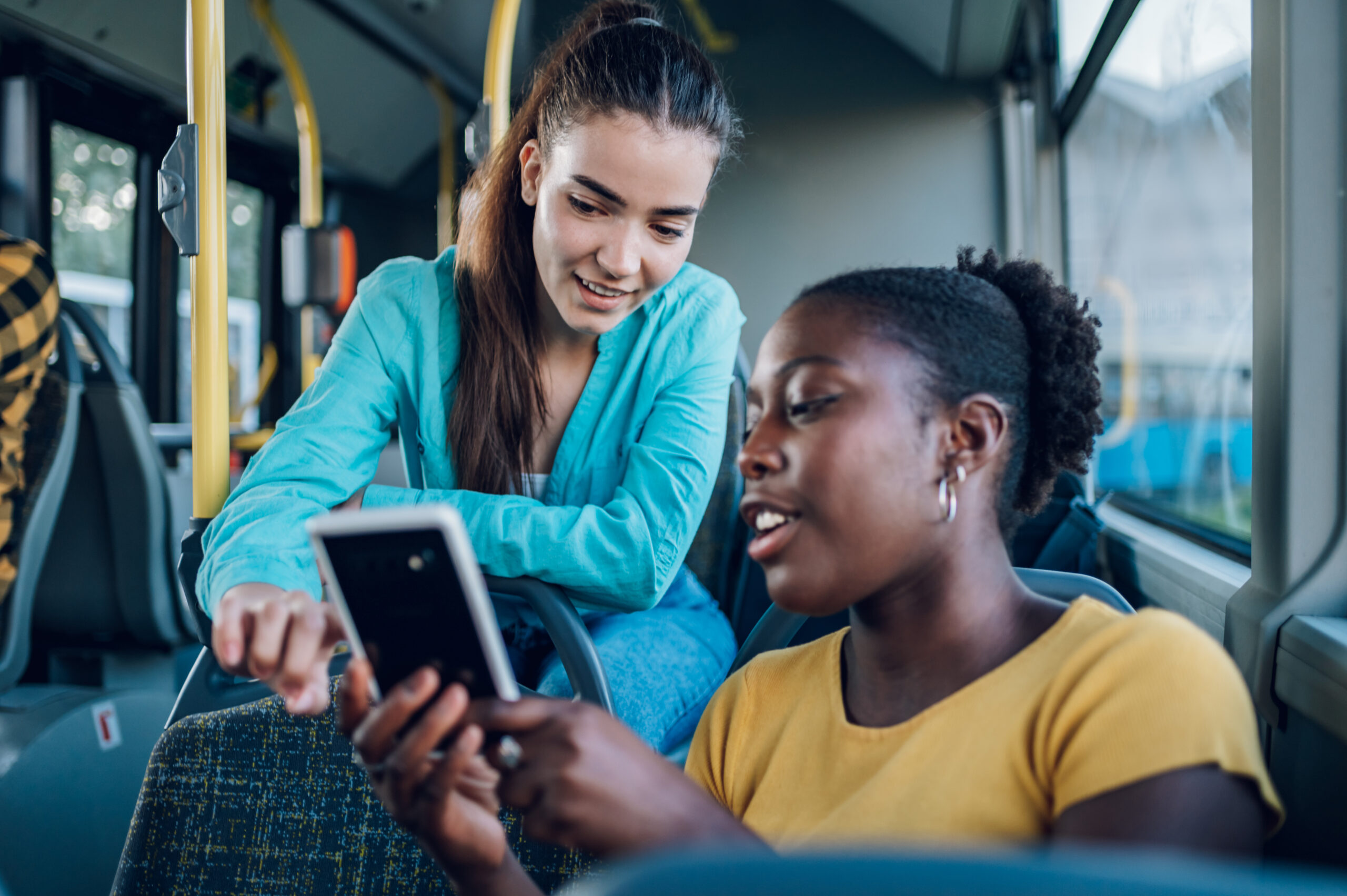 Zwei Frauen lachen gemeinsam und nutzen auf einer Busfahrt ein Smartphone.