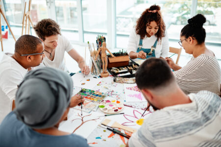 Gruppe von Menschen sitzt gemeinsam an einem Tisch und malt mit Pinseln und Farben in einem hellen Kunstraum. Auf dem Tisch liegen Malutensilien, Paletten und verschiedene Zeichnungen.