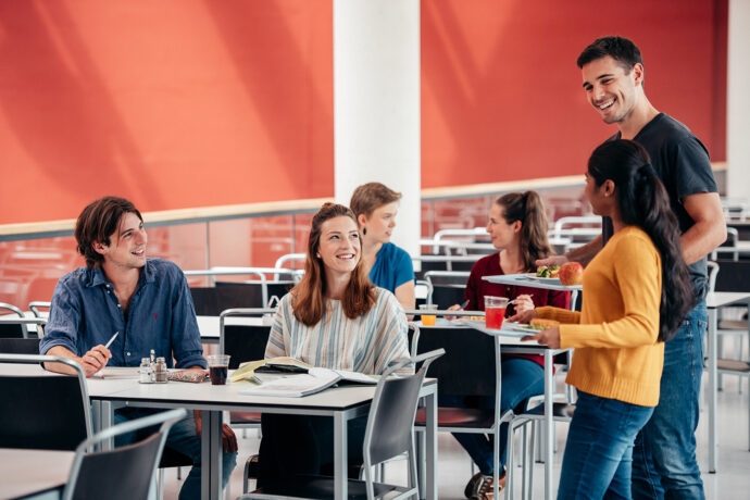 Studierende, die in der Mensa sitzen, sich unterhalten und miteinander lachen. Einige haben ein Tablett mit Mensaessen und Getränken in der Hand.