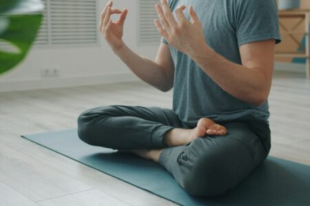 Mensch sitzt im Schneidersitz auf einer Yogamatte, die Hände im Mudra. Sein Gesicht sieht man nicht, nur seinen Körper ab Brusthöhe.