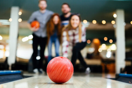 Fokus ist auf einer roten Bowlingkugel, die auf einer Bowlingbahn rollt. Im Hintergrund stehen vier junge Menschen, die unscharf sind.